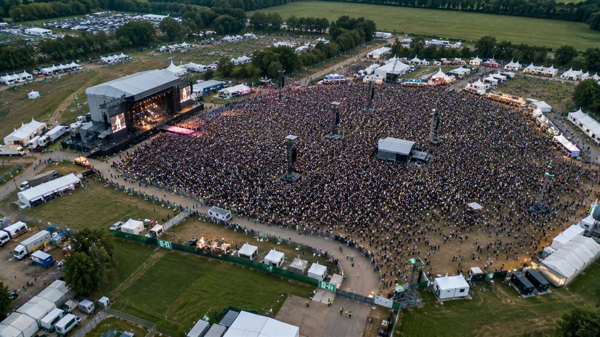Luftaufnahme eines Open-Air-Veranstaltungsgeländes mit tausenden Besuchern vor einer Bühne, umgeben von gekennzeichneten Rettungswegen und Zelten (KI-generiertes Bild)